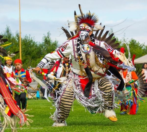 Auf einer Wiese beim Pow Wow in Québec trägt ein Tänzer aus Pessamit-Betsiamites eine auffällige, bunte Federtracht mit Schmuck und Masken. Kanada-Rundreise