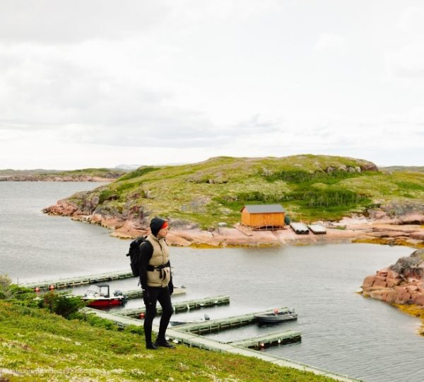 Am Ufer von Tête-a-la-Baleine mit grünen Hügeln, felsigen Küsten und ruhigem Wasser führt ein Steg an einer kleinen Hütte vorbei. Kanada-Rundreise
