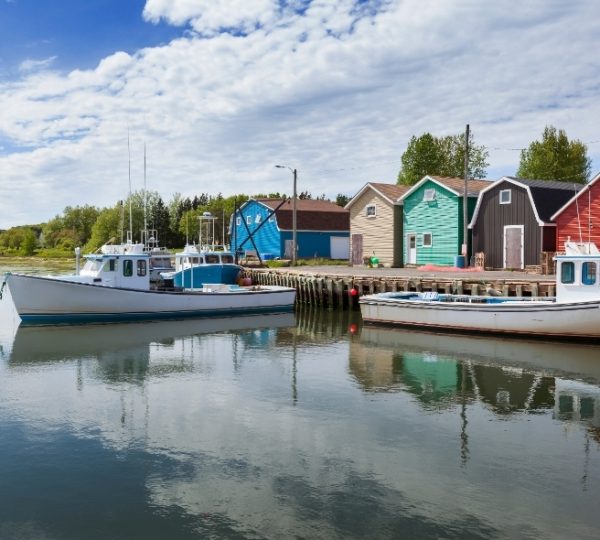 Bunte Holzhäuschen am Ufer von Prince Edward Island, gelbe, grüne und rote Häuser reihen sich am Kai neben zwei weißen Fischerbooten, ruhiges Wasser spiegelt die Szenerie Kanada-Rundreise