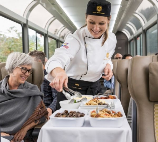 Auf dem Rocky Mountaineer serviert das Küchenteam ein Menü im Zug mit Panoramafenstern, während Reisende bequem an den Sitzen sitzen. Kanada-Rundreise
