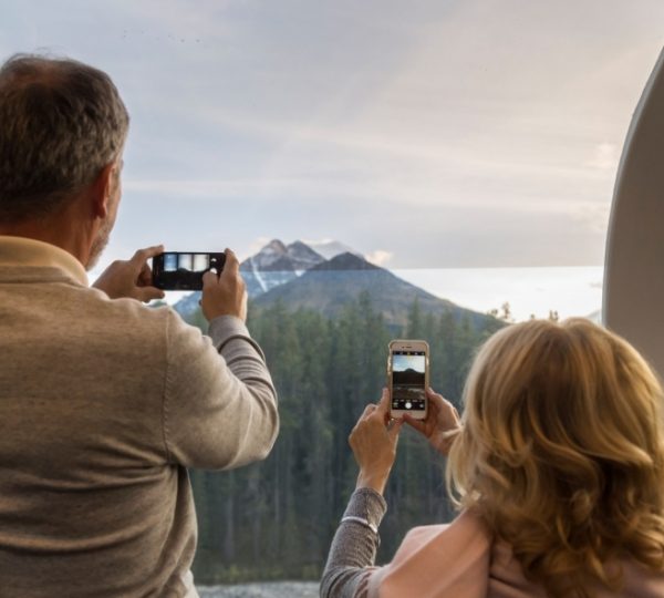 Zwei Reisende halten im Panoramafenster des Rocky Mountaineer mit Smartphones fest, wie sich die Bergkulisse in Westkanada bei Banff zeigt. Kanada-Rundreise