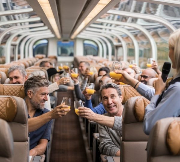 Im Inneren des Rocky Mountaineer Zuges stoßen Reisende mit Getränken an, während beige Sitze und Panoramafenster den Blick in die Landschaft öffnen. Kanada-Rundreise