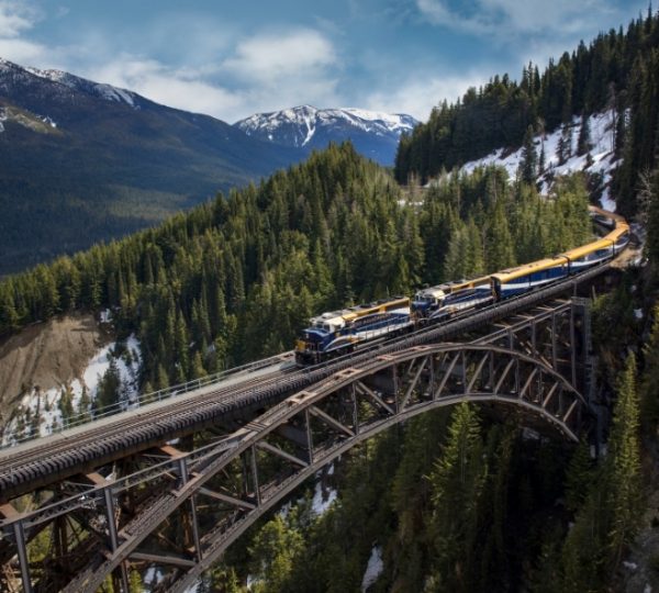 Gelb-blauer Rocky Mountaineer Zug fährt über eine Brücke durch dichte Wälder und verschneite Rocky Mountains in Westkanada während der Kanada Zugreise. Kanada-Rundreise