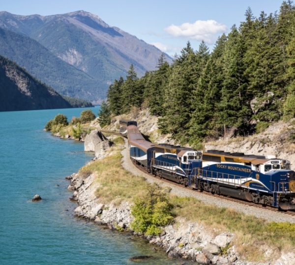 Ein Rocky Mountaineer Zug fährt entlang eines türkisfarbenen Sees durch bewaldete Berge in den Rocky Mountains und schafft Ruhe in Westkanada. Kanada-Rundreise