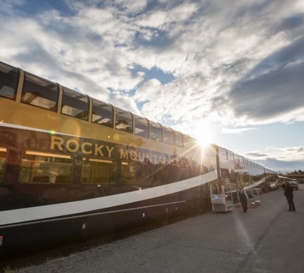 Goldfarbener Rocky Mountaineer Zug mit großen Panoramafenstern an einem Bahnhof, im Gegenlicht vor klaren Bergen und Westkanada-Landschaft rund um Banff. Kanada-Rundreise