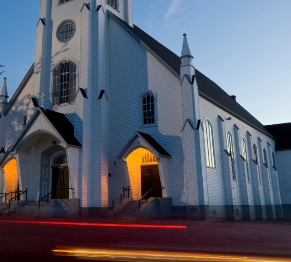 Weiße Kirche in Sydney, Nova Scotia, mit hohen Fenstern und spitzen Türmchen, beleuchtetem Eingang im Abendlicht, ruhige Straßenszene Kanada-Rundreise