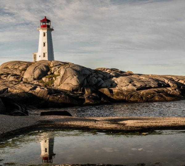 Ein roter Leuchtturm thront auf grauen Felsen am Meeresufer, sanfte Wolken am Himmel, ruhiges Wasser spiegelt den Turm und die Küstenlandschaft von Nova Scotia wider, Sydney Kanada-Rundreise