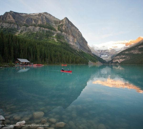 Ruhiger türkisfarbener See bei Lake Louise in Alberta, umgeben von Fels- und schneebedeckten Bergen; rote Kanus gleiten über stilles Wasser. Kanada-Rundreise