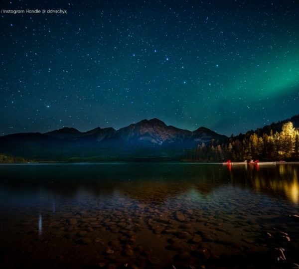Ruhiges Wasser des Pyramid Lake unter sternklarem Himmel im Jasper National Park, Alberta, mit Bergsilhouetten und leuchtendem Nordlicht. Kanada-Rundreise
