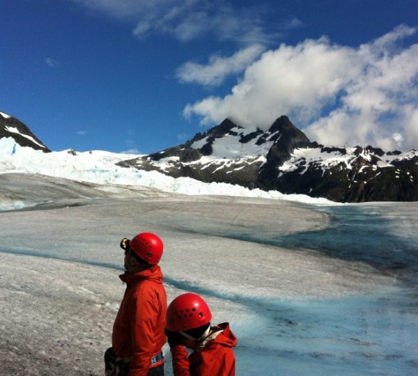 Zwei Bergsteiger in roter Ausrüstung mit Helmen stehen vor einer schneeweißen Gletscherlandschaft in Alaska; dunkle Berggipfel, blaues Wasser und blauer Himmel im Hintergrund Kanada-Rundreise