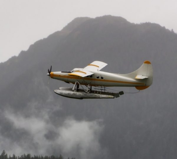 Weißes Schwimmerflugzeug mit orangefarbener Markenführung fliegt über nebeligem Bergmassiv in Alaska, Juneau als Bezugspunkt, Schwimmerboot unter dem Rumpf sichtbar Kanada-Rundreise