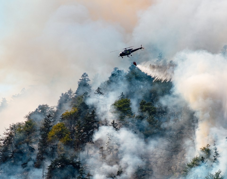Ein Löschhubschrauber wirft Wasser über einen stark verrauchten, bewaldeten Hang im Fraser Canyon in British Columbia ab. Kanada-Rundreise