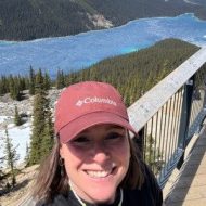 Eine lächelnde Frau mit roter Columbia-Kappe steht auf einer Aussichtsplattform vor einem türkisblauen Bergsee und Nadelwald. Kanada-Rundreise
