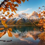 Ein Bergsee spiegelt schneebedeckte Gipfel, während orangefarbenes Herbstlaub den Indian Summer in Kanada rahmt. Kanada-Rundreise
