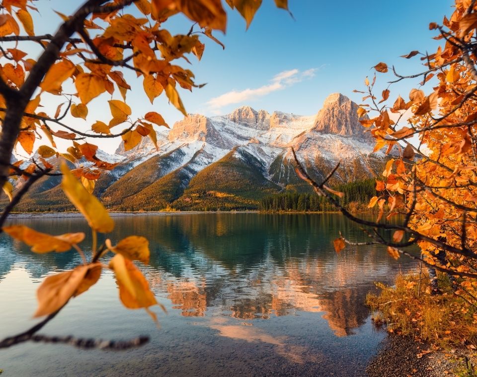 Ein Bergsee spiegelt schneebedeckte Gipfel, während orangefarbenes Herbstlaub den Indian Summer in Kanada rahmt. Kanada-Rundreise