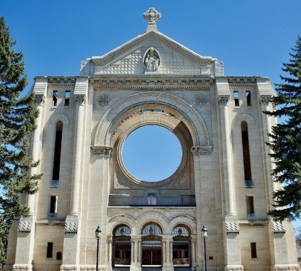 Historische helle Kirche mit großem Rundbogen und Kreuz zwischen hohen Nadelbäumen in Churchill, Kanada Kanada-Rundreise